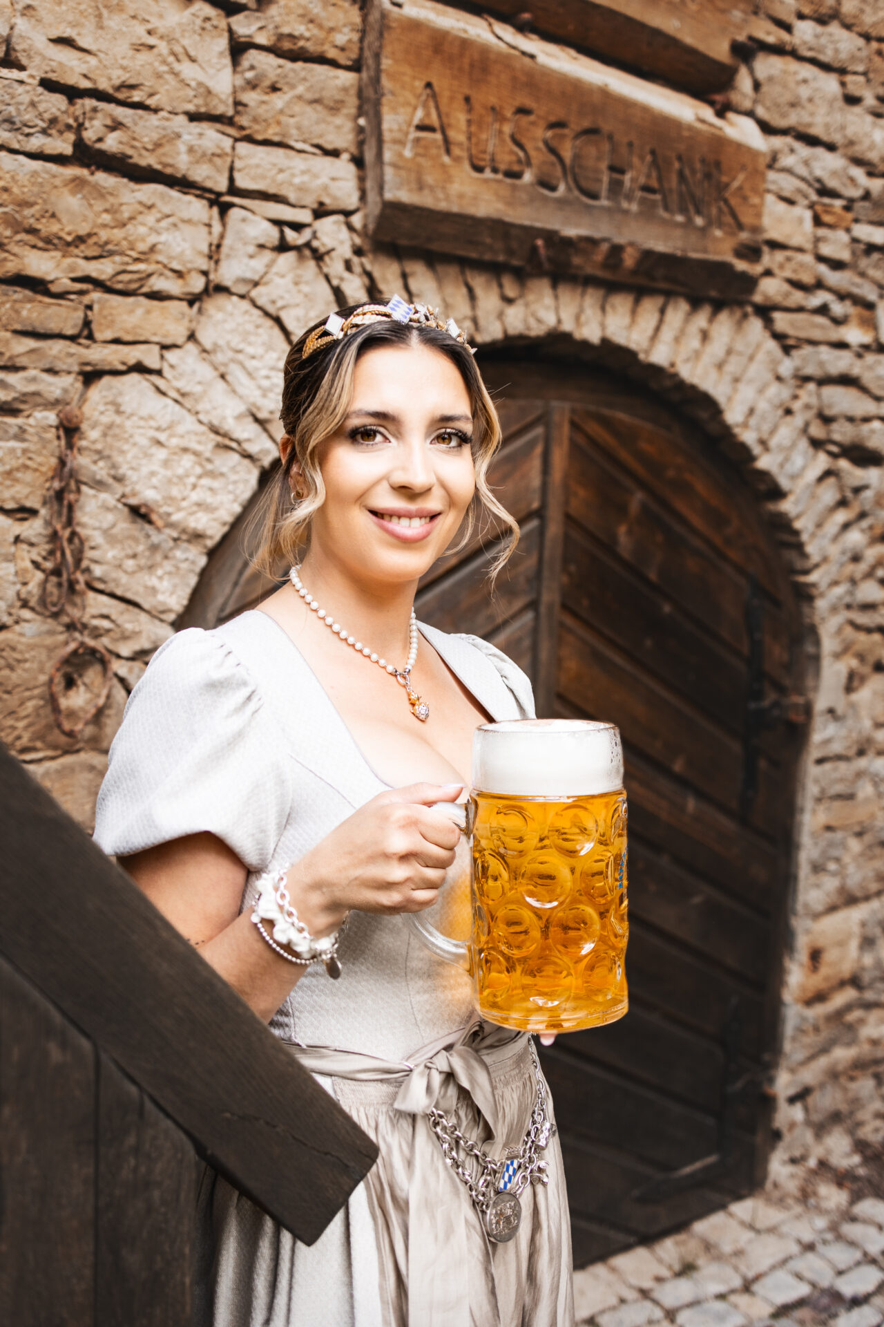 Junge, schöne Frau im Dirndl mit Krone und einem frisch eingeschenkten Maßkrug. Sie lächelt in die Kamera.