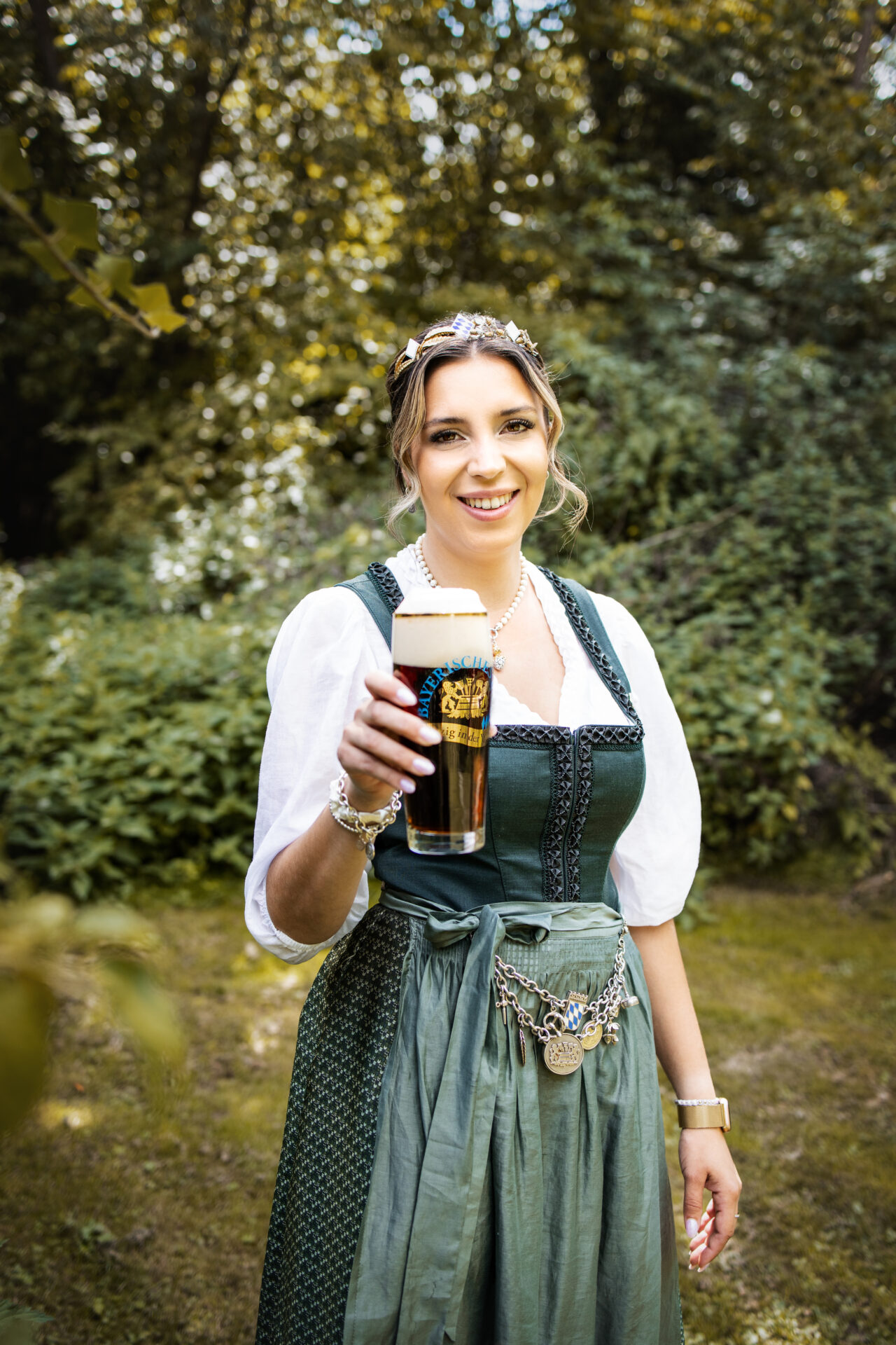 Junge, schöne Frau im Dirndl mit Krone und einem frisch dunklen Bier. Sie lächelt in die Kamera.